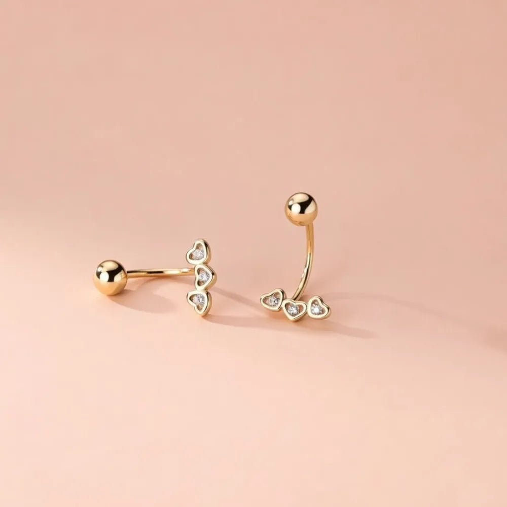 Goldtonade navelpiercingar med tre hjärtformade berlocker och klara kristaller från ari örhängen med krokdesign och skruvplopp