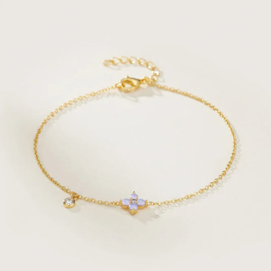 Azuralie armband med en tunn guldkedja och ett litet lavendelfärgat fyra-petal blomma-berlock med klar sten