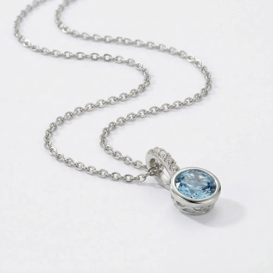 A delicate silver chain necklace with a round light blue gemstone pendant and filigree detailing called blue ivy